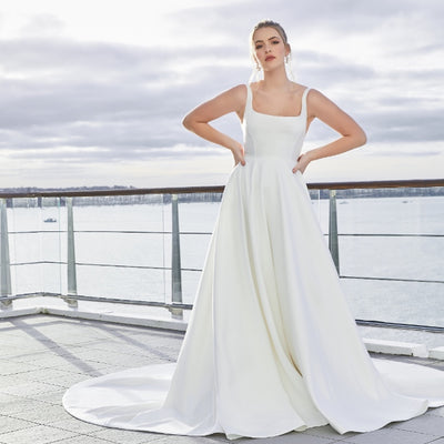 Minimalist ivory satin a-line gown with square neckline. Trinity has a dramatic skirt with pockets and a long train.