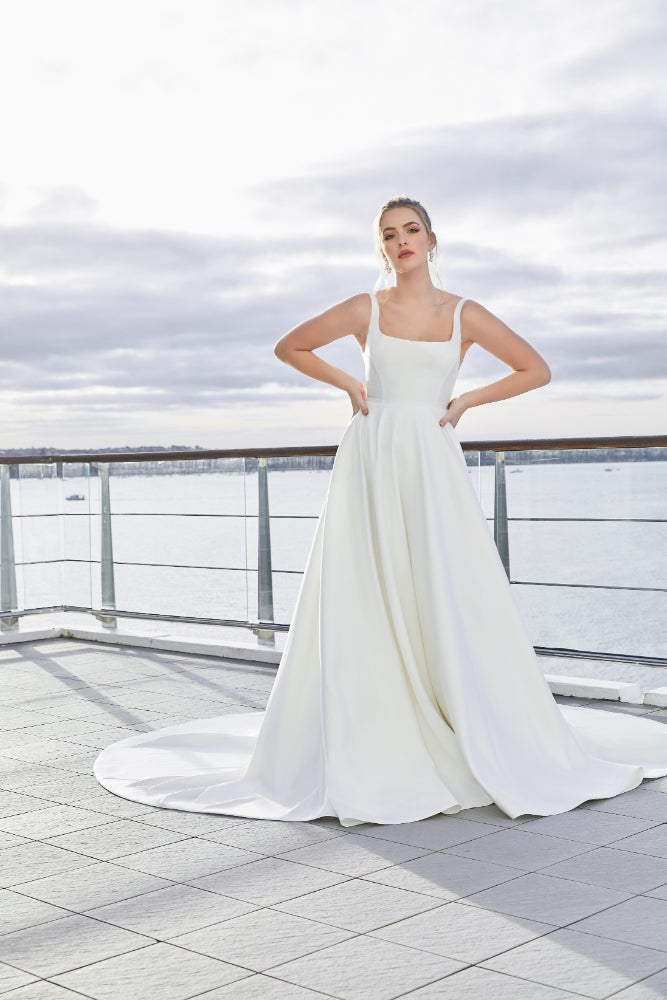 Minimalist ivory satin a-line gown with square neckline. Trinity has a dramatic skirt with pockets and a long train.