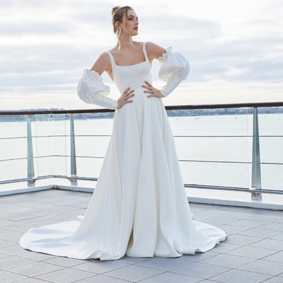 Minimalist ivory satin a-line gown with square neckline. Trinity has a dramatic skirt with pockets. Pictured here with detached balloon sleeves with elbow length cuffs with covered buttons