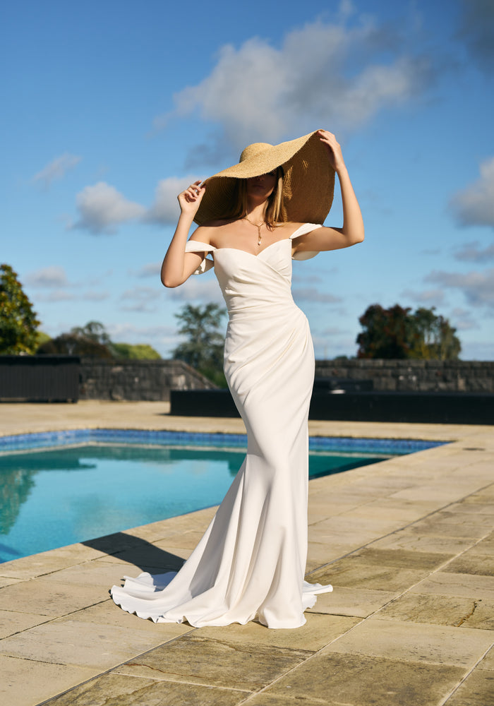 Priscilla gown in ivory crepe. Ruched crossover fit-n-flare silhouette with off-shoulder draped straps.