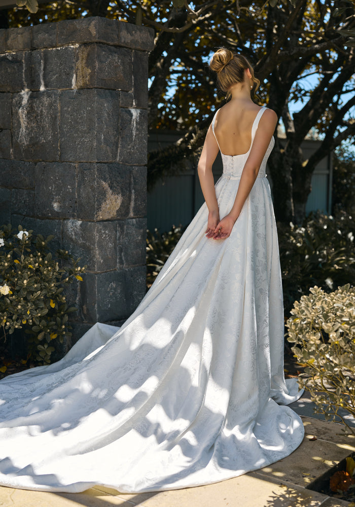 Model wears Perry gown with detachable full a-line overskirt. Floral jacquard fabric and scoop back.